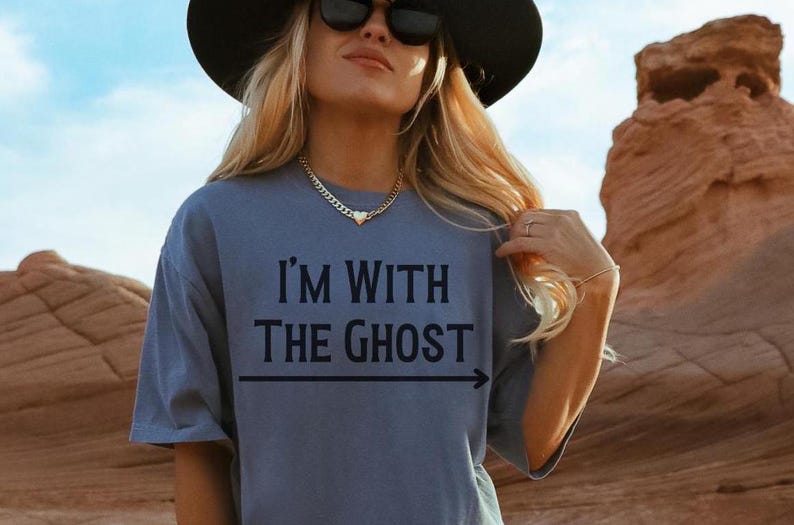 May include: A woman wearing a blue t-shirt with the text "I'm with the ghost" and an arrow pointing to the right. She is wearing a black hat and sunglasses. The background is a desert landscape.