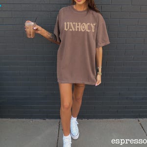 Op de afbeelding: Een vrouw draagt een oversized bruin t-shirt met het woord "UNHOLY" erop gedrukt in een bruin lettertype. Ze draagt witte sneakers en houdt een kop koffie vast.
