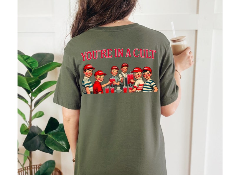 K&ouml;nnte beinhalten: Ein gr&uuml;nes T-Shirt mit dem Text "YOU'RE IN A CULT" auf der R&uuml;ckseite. Der Text ist rot. Das Shirt zeigt eine Vintage-Illustration einer Gruppe von Jungen, die aus einem Brunnen trinken.