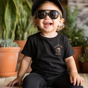 May include: A young child wearing a black t-shirt with the text "Rad Like My Dad" and a skull graphic. The child is also wearing a black cap and sunglasses and is sitting on a woven rug.