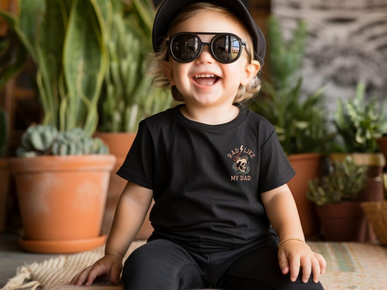 May include: A young child wearing a black t-shirt with the text "Rad Like My Dad" and a skull graphic. The child is also wearing a black cap and sunglasses and is sitting on a woven rug.