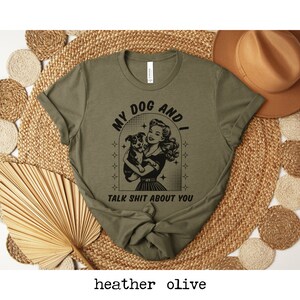 Op de afbeelding: Een groen T-shirt met een zwarte afbeelding van een vrouw die een hond vasthoudt. De afbeelding zegt "My Dog And I Talk Shit About You".