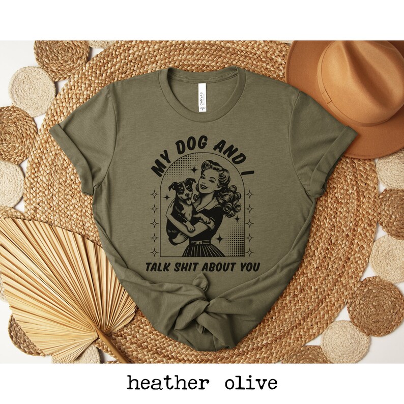 Op de afbeelding: Een groen T-shirt met een zwarte afbeelding van een vrouw die een hond vasthoudt. De afbeelding zegt "My Dog And I Talk Shit About You".