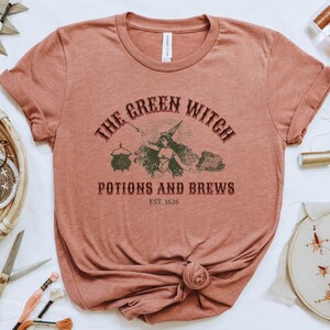 May include: A rust-coloured t-shirt with a vintage-style graphic that reads "The Green Witch Potions and Brews Est. 1626". The graphic features a witch with a cauldron and other potion-making supplies.