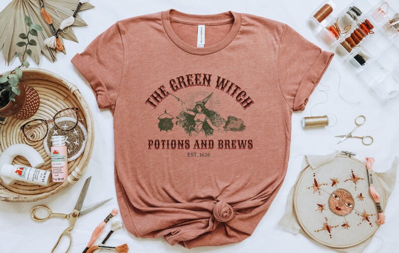 May include: A rust-coloured t-shirt with a vintage-style graphic that reads "The Green Witch Potions and Brews Est. 1626". The graphic features a witch with a cauldron and other potion-making supplies.
