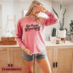 May include: A pink long-sleeved t-shirt with the text "Go Sage Yourself" in a retro font. The shirt is made of a soft, comfortable material and has a relaxed fit.