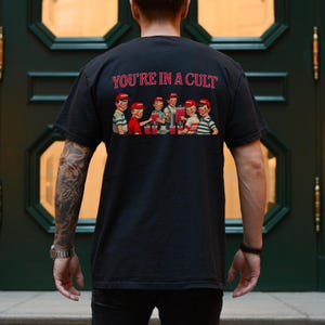 K&ouml;nnte beinhalten: Ein schwarzes T-Shirt mit einem grafischen Druck einer Gruppe von Kindern, die aus einem gro&szlig;en Krug trinken. Der Text "YOU'RE IN A CULT" ist &uuml;ber dem Grafikdruck gedruckt.