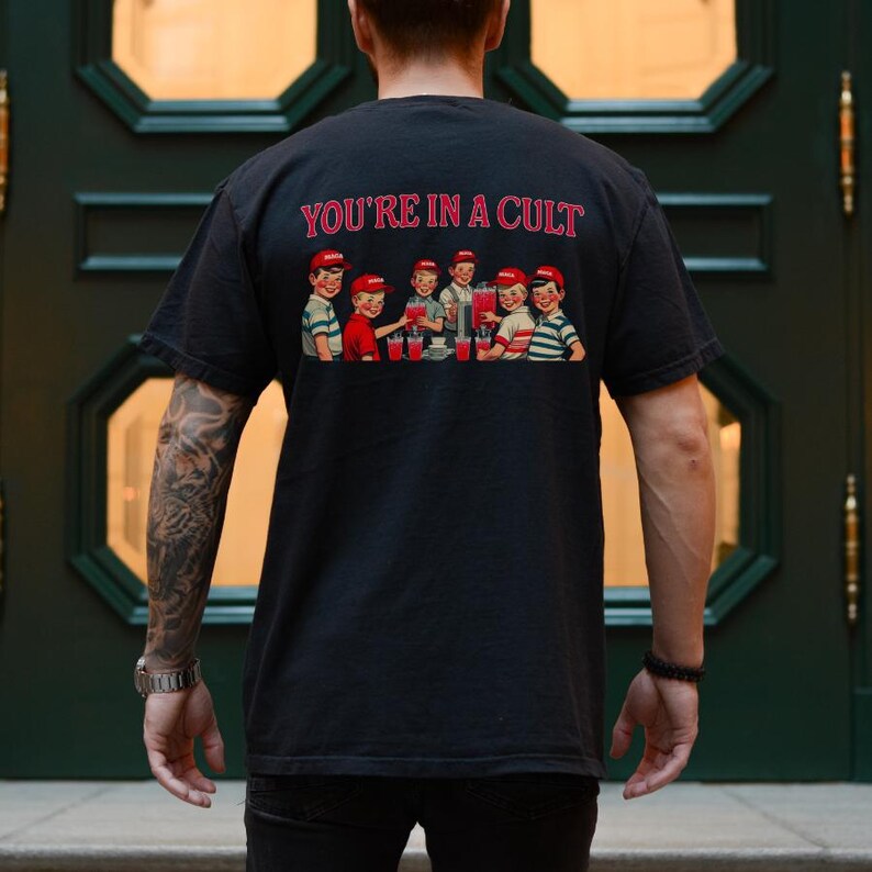 K&ouml;nnte beinhalten: Ein schwarzes T-Shirt mit einem grafischen Druck einer Gruppe von Kindern, die aus einem gro&szlig;en Krug trinken. Der Text "YOU'RE IN A CULT" ist &uuml;ber dem Grafikdruck gedruckt.