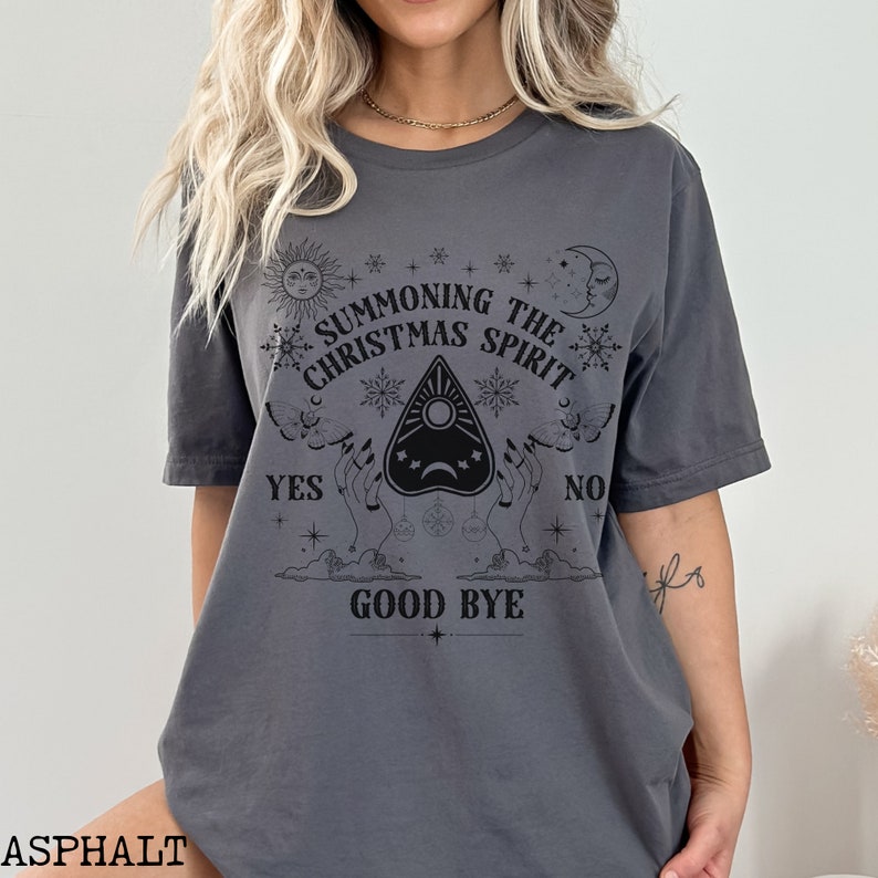 May include: A gray t-shirt with a black graphic design that says "Summoning the Christmas Spirit". The graphic includes a Ouija board planchette, stars, moons, and the words "Yes", "No", and "Good Bye".
