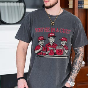 May include: A dark gray t-shirt with a graphic print of three children wearing red hats and holding pitchers of red liquid. The text "YOU'RE IN A CULT" is printed above the graphic. The children's hats have the word "MAGA" printed on them.