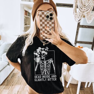 May include: Black t-shirt with a skeleton graphic holding a cup of coffee. The text "Dead inside and slightly bitter" is printed below the graphic.