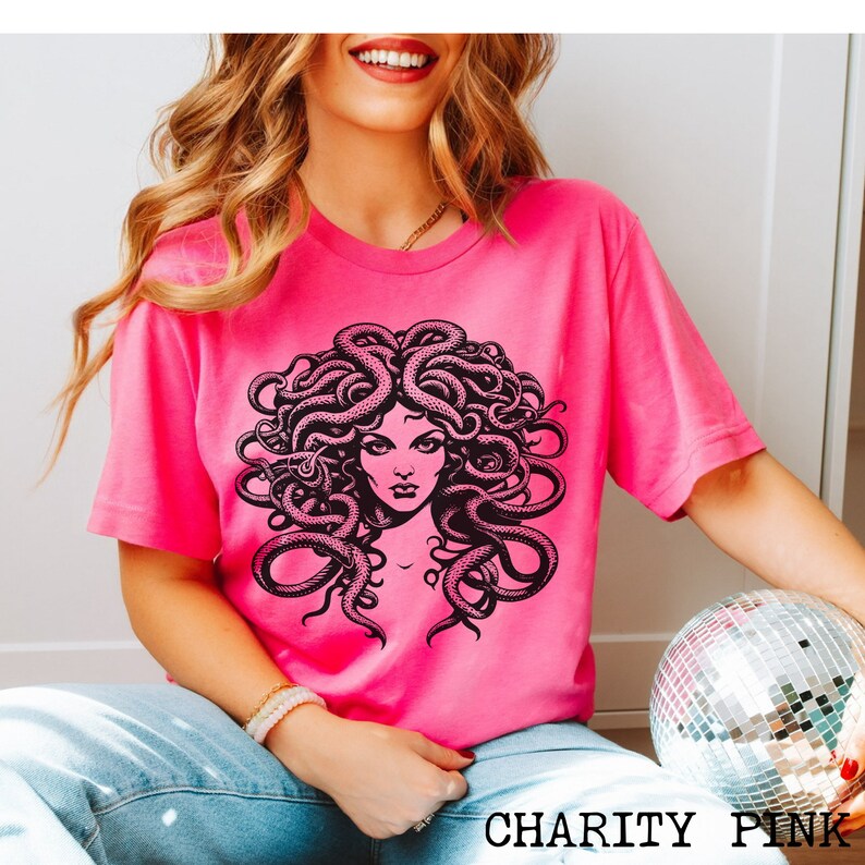 May include: A pink t-shirt with a black and white graphic of Medusa, a mythical Greek figure with snakes for hair. The text "CHARITY PINK" is printed on the bottom of the shirt.