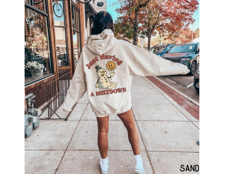 May include: Sand-colored hoodie with a graphic on the back that reads "Just Having A Meltdown" with a melting snowman and smiling sun. The hoodie is oversized and has a hood.