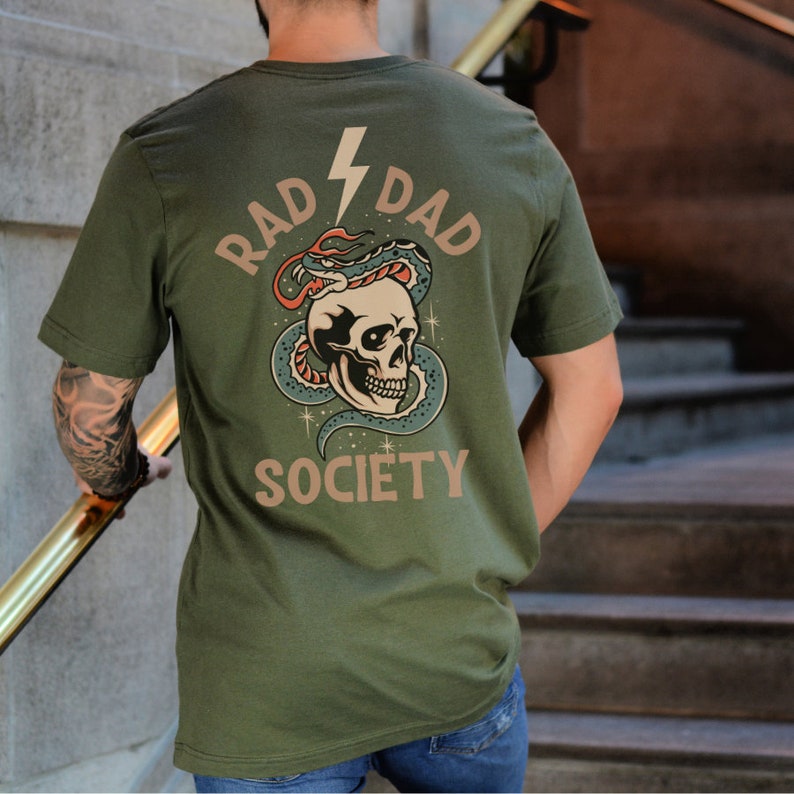 May include: A green t-shirt with a graphic of a skull with a snake wrapped around it. The text "RAD DAD SOCIETY" is printed on the shirt.