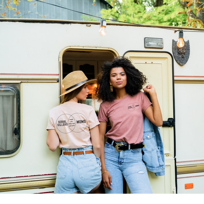Puede incluir: Dos mujeres con camisetas con un gr&aacute;fico de diagrama de Venn que dice "Serial Killers" y "Moms". La mujer de la izquierda lleva una camiseta beige claro y la mujer de la derecha lleva una camiseta rosa. Est&aacute;n de pie frente a una autocaravana blanca.