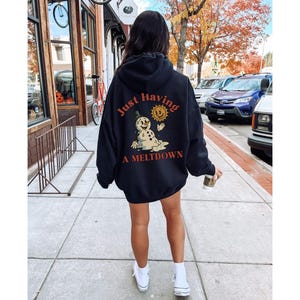 May include: Black hooded sweatshirt with the text "Just Having A Meltdown" and a graphic of a melting snowman and sun. The sweatshirt is worn by a person walking on a sidewalk.