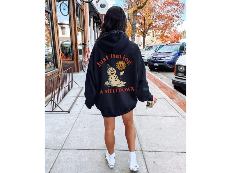 May include: Black hooded sweatshirt with the text "Just Having A Meltdown" and a graphic of a melting snowman and sun. The sweatshirt is worn by a person walking on a sidewalk.