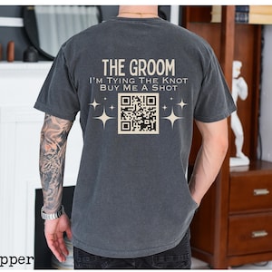 Op de afbeelding: Donkergrijs t-shirt met de tekst "THE GROOM" en "I'M TYING THE KNOT BUY ME A SHOT" op de achterkant. Voorzien van een QR-code en sterrenburst graphics. Een casual t-shirt voor een bruiloft.
