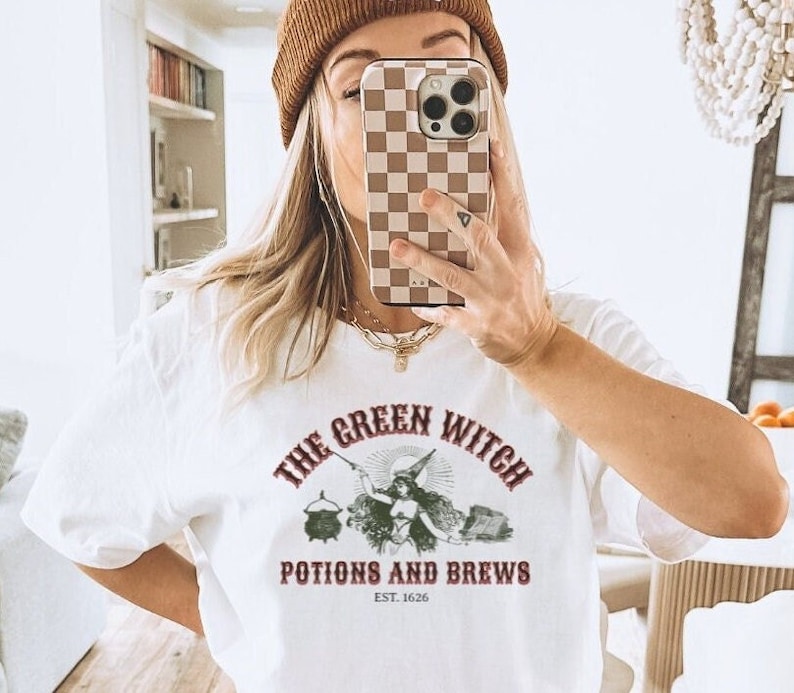 May include: White t-shirt featuring a green witch graphic and the text "The Green Witch Potions and Brews Est. 1626".