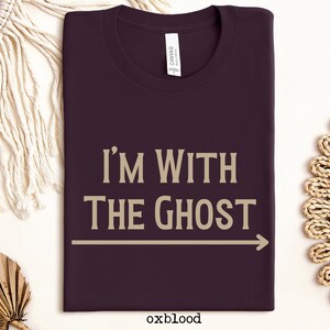 May include: A dark purple t-shirt with the text "I'm With The Ghost" in a light brown color. An arrow points to the right.