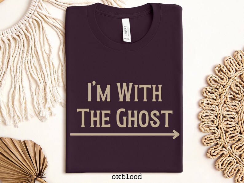 May include: A dark purple t-shirt with the text "I'm With The Ghost" in a light brown color. An arrow points to the right.