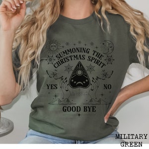 May include: A green t-shirt with a black graphic design that says "Summoning the Christmas Spirit" with a Ouija board design and the words "Yes", "No", and "Good Bye".