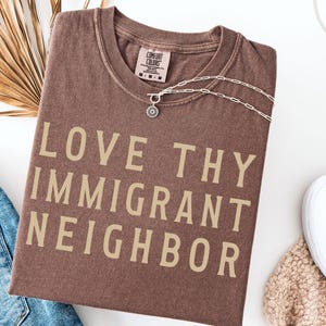 May include: A brown t-shirt with the text "LOVE THY IMMIGRANT NEIGHBOR" in tan lettering. A silver necklace with a circular pendant is on the shirt. The shirt is folded on a white surface, with blue jeans and white shoes visible.