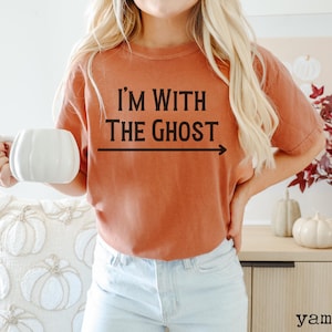 May include: A woman wearing a burnt orange t-shirt with the text "I'm with the ghost" and an arrow pointing to the right. She is holding a white mug and wearing light blue jeans.