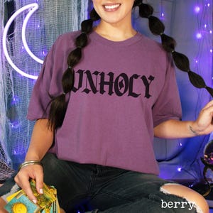 Op de afbeelding: Een paars T-shirt met het woord "UNHOLY" gedrukt in zwarte gotische letters.