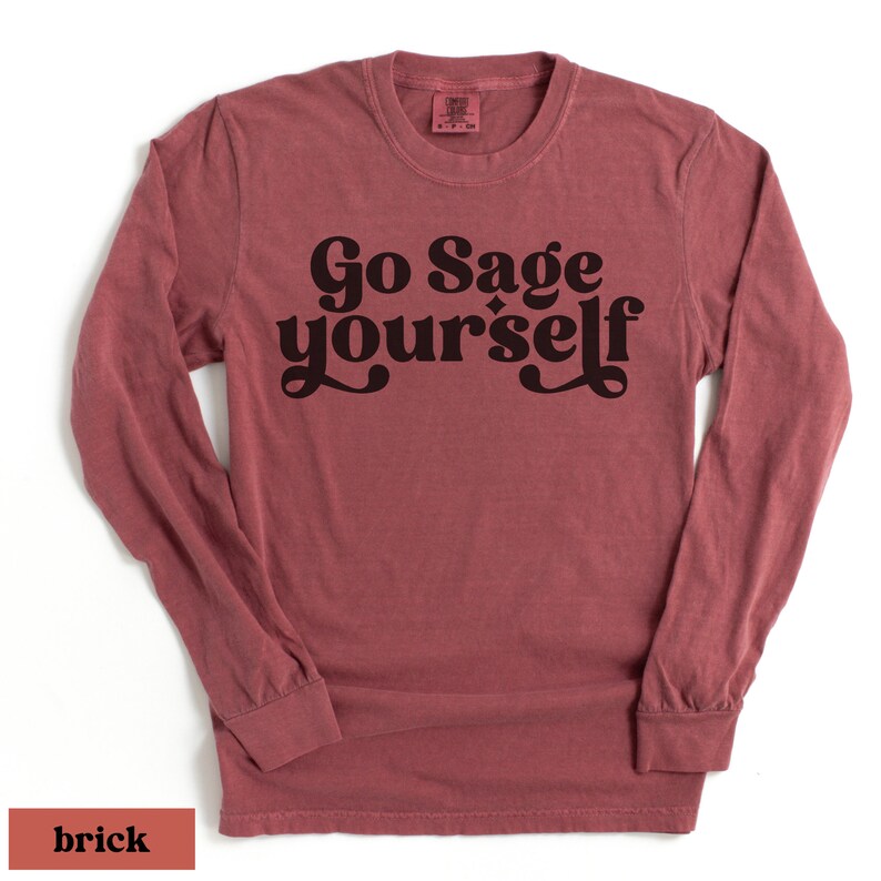 May include: A long-sleeved t-shirt in a brick red color with the text "Go Sage Yourself" printed in black.