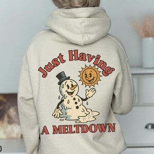 May include: Sand-colored hoodie with a graphic on the back featuring a melting snowman wearing a top hat, a smiling sun, and the text "Just Having A Meltdown" in red. The hoodie is a neutral color and has a hood.