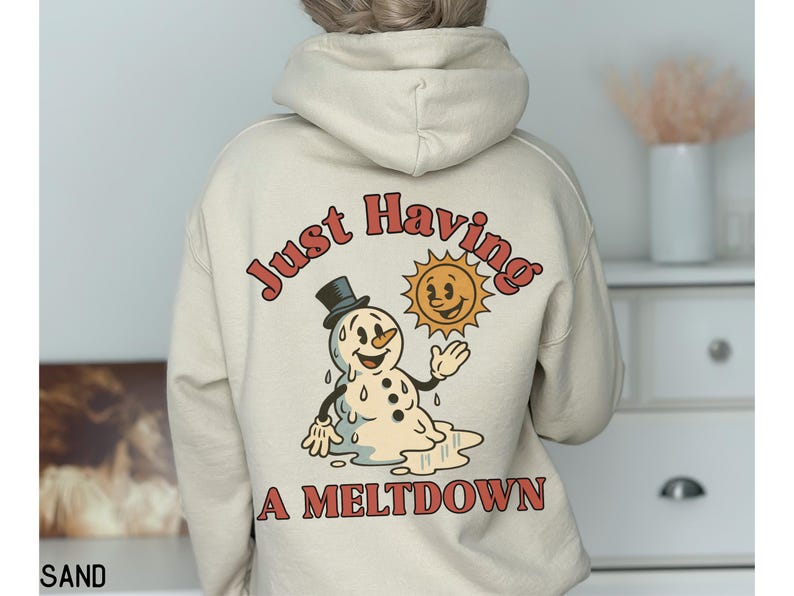 May include: Sand-colored hoodie with a graphic on the back featuring a melting snowman wearing a top hat, a smiling sun, and the text "Just Having A Meltdown" in red. The hoodie is a neutral color and has a hood.