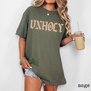 Op de afbeelding: Oversized T-shirt in saliegroen met het woord "UNHOLY" gedrukt in een bruin gotisch lettertype.