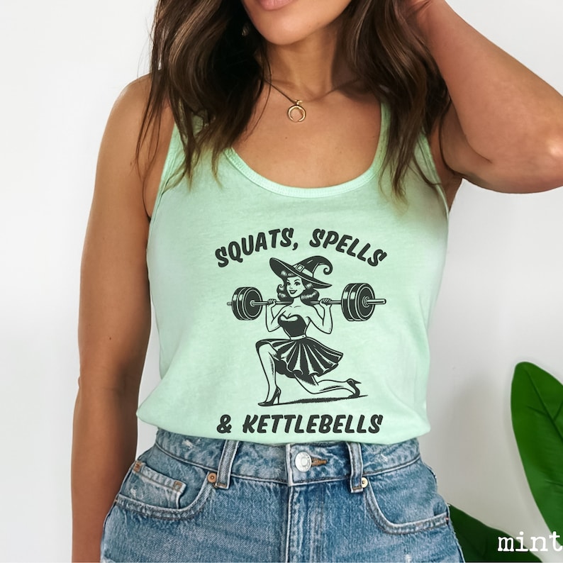 May include: A light green tank top with a black graphic of a witch lifting a barbell. The text on the tank top reads "SQUATS, SPELLS & KETTLEBELLS".