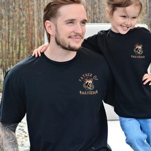 以下が含まれることがあります： 父親と娘が、骸骨のグラフィックと「Father of Mayhem」と「Mayhem」の文字が入ったお揃いの黒いTシャツを着ています。