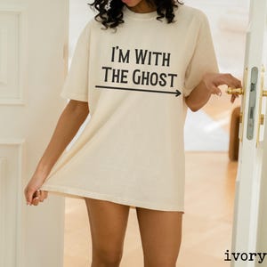 May include: A white oversized t-shirt with the text "I'm with the ghost" and an arrow pointing to the right. The shirt is being worn by a person.