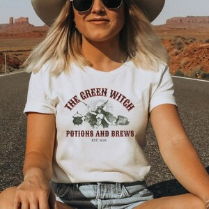 May include: A white t-shirt featuring a vintage-style graphic of a witch holding a wand and a cauldron. The text reads "The Green Witch Potions and Brews Est. 1626".