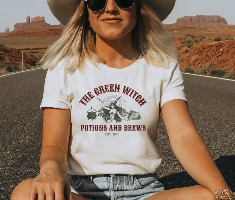 May include: A white t-shirt featuring a vintage-style graphic of a witch holding a wand and a cauldron. The text reads "The Green Witch Potions and Brews Est. 1626".
