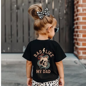 May include: A black t-shirt with a graphic of a skull with a snake wrapped around it. The text "Rad Like My Dad" is printed on the shirt.