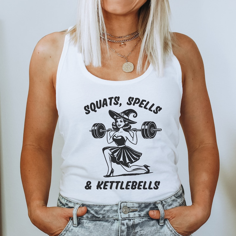 May include: White tank top with a black and white illustration of a witch lifting a barbell. The text on the shirt reads "Squats, Spells & Kettlebells".