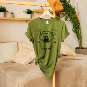 May include: A green t-shirt with a black graphic design that says "Summoning the Christmas Spirit" with a Ouija board graphic and the words "Yes", "No", and "Good Bye".