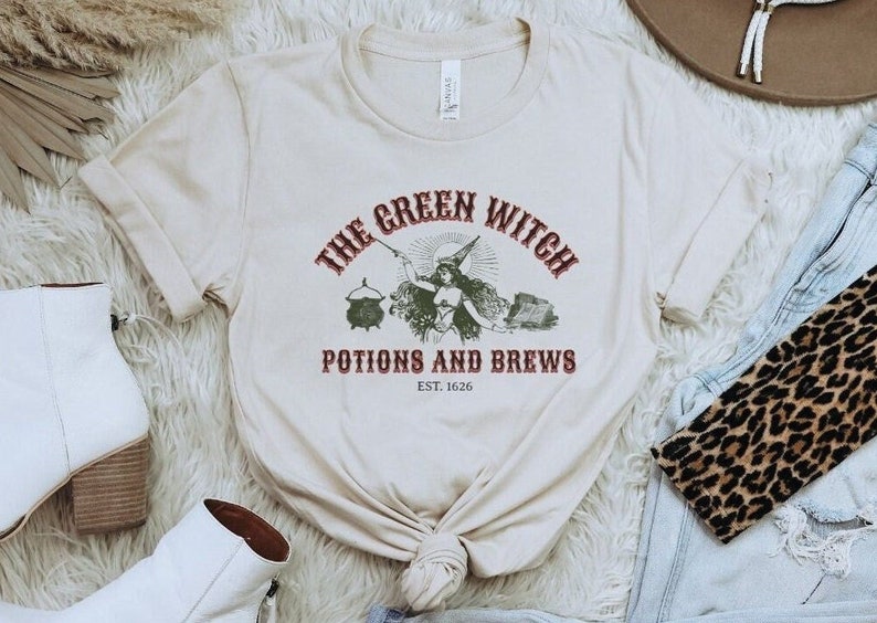 May include: A beige t-shirt featuring a vintage-style graphic of a witch holding a cauldron and a wand. The text reads "The Green Witch Potions and Brews Est. 1626".