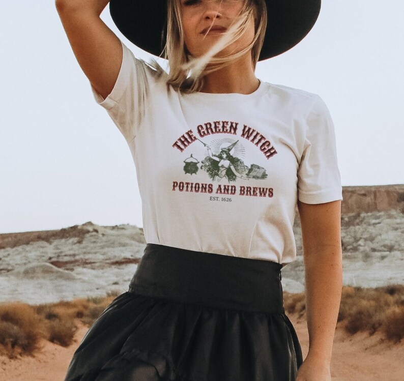 May include: White t-shirt with a green witch graphic and the text "The Green Witch Potions and Brews Est. 1626".