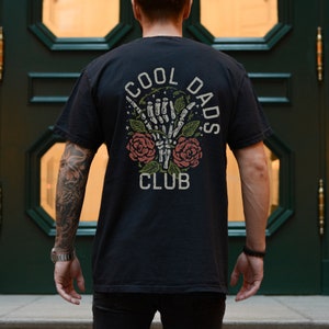 May include: Black t-shirt with a graphic print of a skeleton hand giving a shaka sign with roses and vines. The text "Cool Dads Club" is printed on the shirt.