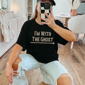 May include: A black t-shirt with the text "I'm with the ghost" in beige lettering and an arrow pointing to the right. The shirt is being worn by a person with light brown hair and a black baseball cap.