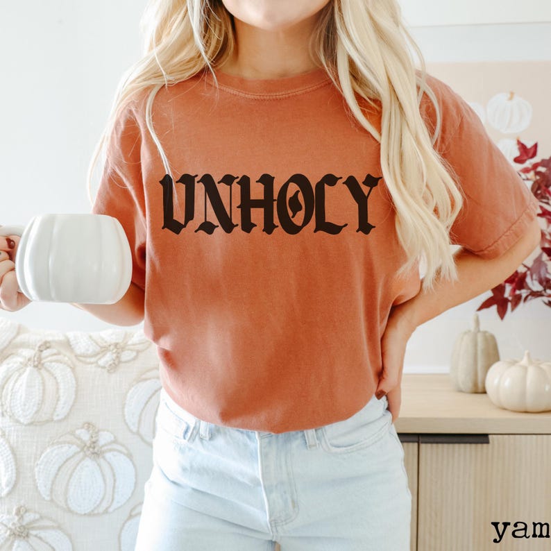Op de afbeelding: Een vrouw draagt een oranje t-shirt met het woord "UNHOLY" gedrukt in zwarte gotische letters. Ze draagt lichtblauwe jeans en houdt een witte mok vast.