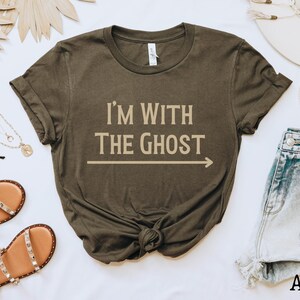 May include: A dark green t-shirt with the text "I'm with the Ghost" in beige lettering and an arrow pointing to the right. The shirt is made of a soft, comfortable material.