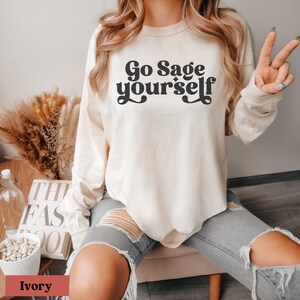 May include: A long-sleeved, ivory-colored t-shirt with the text "Go Sage yourself" printed in black. The text is in a retro font style.