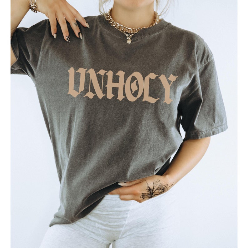 Op de afbeelding: Een donkergrijs T-shirt met het woord "UNHOLY" gedrukt in een bruin gotisch lettertype.