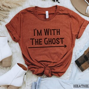 May include: A rust-colored t-shirt with the text "I'm with the ghost" and an arrow pointing to the right. The shirt is folded and displayed on a white surface.